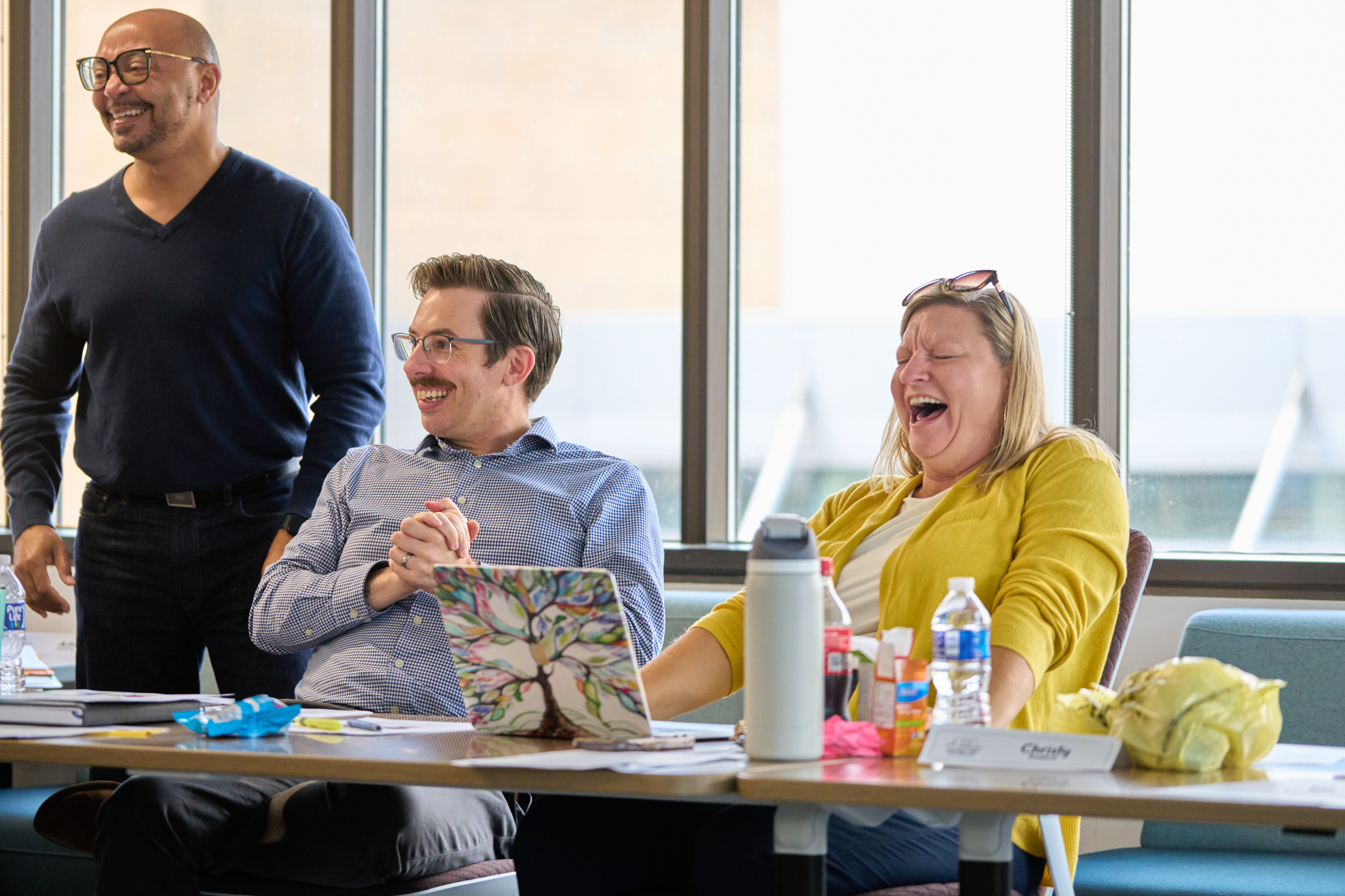 Three people smiling and laughing. Two sitting. One standing. 